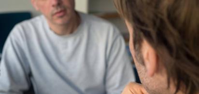 Zwei männlich gelesene Personen sitzen sich in einem ernsten Gespräch gegenüber.