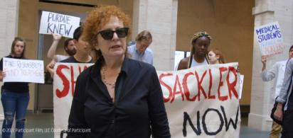Nan Goldin bei einer Protrestaktion mit P.A.I.N: Die Fotografin im schwarzen Hemd mit Sonnenbrille im Vordergrund, dahinter mehrere Personen mit Transparenten