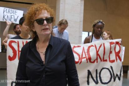 Nan Goldin bei einer Protrestaktion mit P.A.I.N: Die Fotografin im schwarzen Hemd mit Sonnenbrille im Vordergrund, dahinter mehrere Personen mit Transparenten